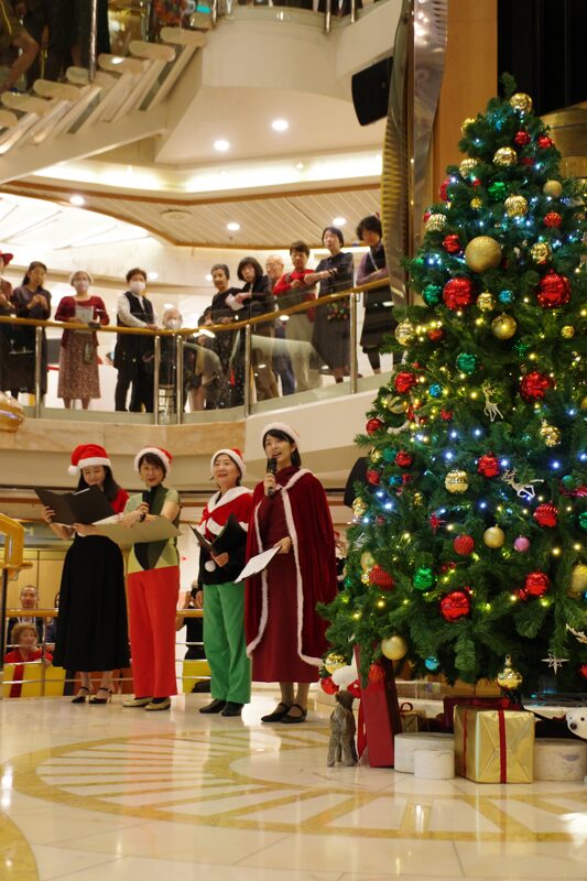 船内で行われたクリスマスパーティ。ツリー横には、CC（コミュニケーション・コーディネーター）が並んだ。主だったイベントは、日本語、中国語、韓国語、英語の4か国語で対応している。